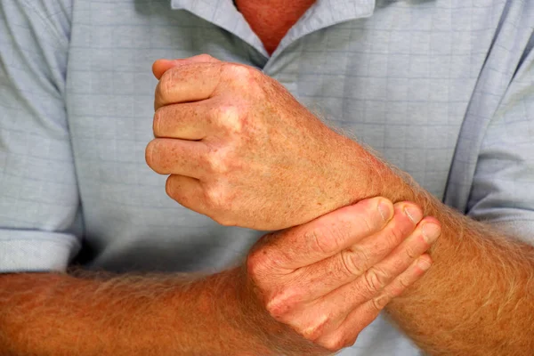 Left wrist of a mature white man wearing a blue shirt being massaged by his right human hand fingers close-up. Close-up of an adult caucasian male massaging his left wrist with his hand and fingers