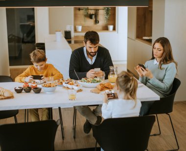 Aile, evdeki yemek masasında kahvaltı ederken cep telefonu kullanıyor.