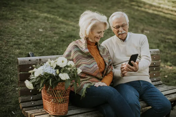 Красивая Пожилая Пара Сидит Скамейке Корзиной Цветов Смотрит Мобильный Телефон — стоковое фото
