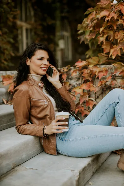 秋の日に屋外の階段に行くために携帯電話のドリンクコーヒーを持つかなり若い女性 — ストック写真