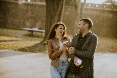 Güz parkında ellerinde hazır kahveyle yürüyen yakışıklı bir çift.