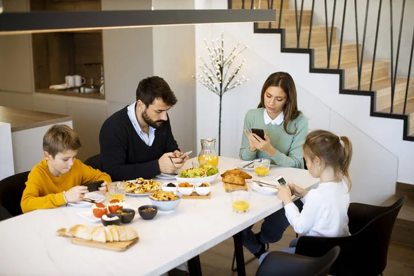 アパートのダイニングテーブルで朝食をとりながら携帯電話を使う家族 — ストック写真
