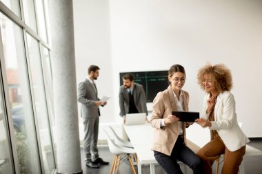 Ofiste dijital tabletle konuşan iki genç iş kadını arkalarında genç insanlarla çalışıyor.