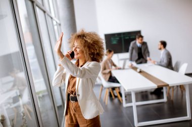 Genç kıvırcık saçlı iş kadını ofiste cep telefonu kullanıyor ve arkasında genç insanlar çalışıyor.