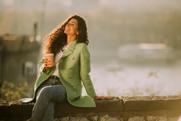 川のそばに座り テイクアウトコーヒーを飲みながら秋の太陽を楽しむ可愛い若い巻き毛の女性 — ストック写真