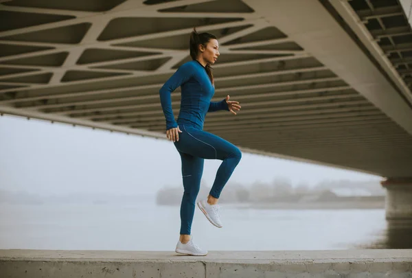 秋天的早晨 身穿蓝色运动服的年轻漂亮女子在河边锻炼前伸展着身体 — 图库照片