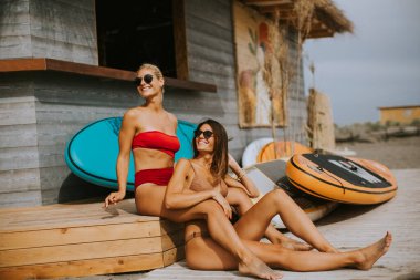 Bikinili güzel kadınlar yaz günü sahilde sörf kulübesinde oturuyor.