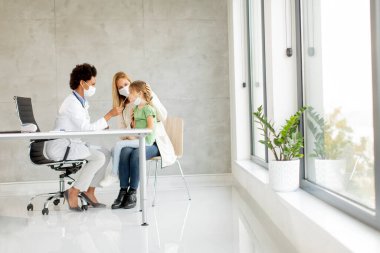 Annesi, küçük kızıyla birlikte pediatri muayenesinde ve Afrikalı Amerikalı kadın doktor tarafından PCR testi yaptırıyor.