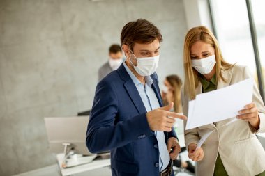 Koruyucu maskeli genç bir adam ve kadın ellerinde kağıtlarla ofisin içinde genç insanlarla tartışıyorlar.
