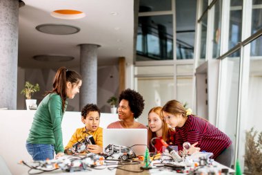 Bir grup mutlu çocuk ve dizüstü bilgisayarı olan Afrika kökenli Amerikalı kadın fen öğretmenleri robot sınıfında elektrikli oyuncaklar ve robotlar programlıyorlar.