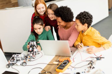 Bir grup mutlu çocuk ve dizüstü bilgisayarı olan Afrika kökenli Amerikalı kadın fen öğretmenleri robot sınıfında elektrikli oyuncaklar ve robotlar programlıyorlar.