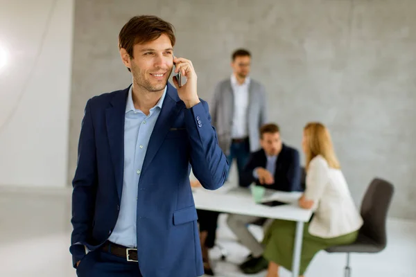彼のチームの前でオフィスに自信を持って立って携帯電話を使用しているハンサムな若いビジネスマン — ストック写真