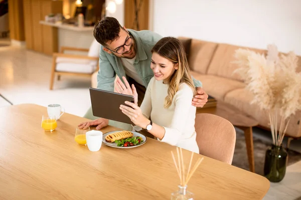 デジタルタブレットを使用して素敵な若いカップルとキッチンで朝食を持っています — ストック写真