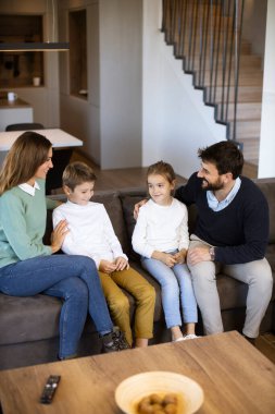 Oturma odasındaki kanepede birlikte televizyon izleyen gülümseyen genç aile.