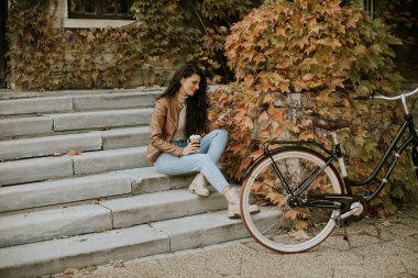 Cep telefonlu güzel bir genç bayan sonbahar günü bisikletle merdivenlerde kahve içer.