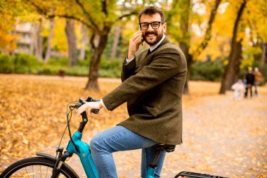 Güz parkında elektrikli bisikletle cep telefonu kullanan yakışıklı genç adam.