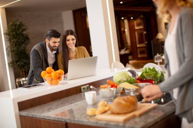 Bir grup mutlu genç yemek hazırlıyor, laptopta yemek tarifleri arıyor ve iyi vakit geçiriyorlar.