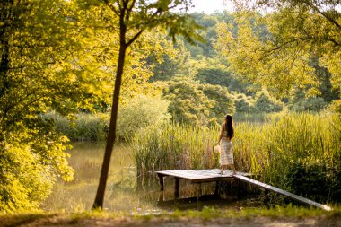Genç bir kadın sıcak bir yaz gününde sakin gölün oradaki ahşap iskelede yürüyor.
