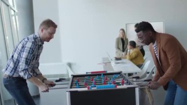 Genç, yakışıklı, çok ırklı iş adamları masa futbolu oynuyor ve ofiste dinleniyor.
