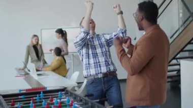 Genç, yakışıklı, çok ırklı iş adamları masa futbolu oynuyor ve ofiste dinleniyor.