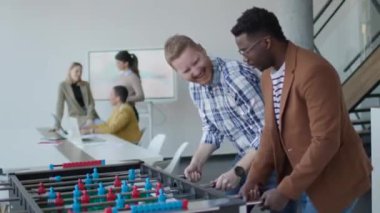 Genç, yakışıklı, çok ırklı iş adamları masa futbolu oynuyor ve ofiste dinleniyor.