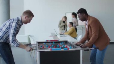 Genç, yakışıklı, çok ırklı iş adamları masa futbolu oynuyor ve ofiste dinleniyor.
