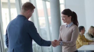 Genç iş ortakları ofiste tokalaşırken takımları arka planda çalışıyor.