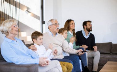 Modern evdeki kanepeye oturup televizyon izleyen çok nesilli bir aile.