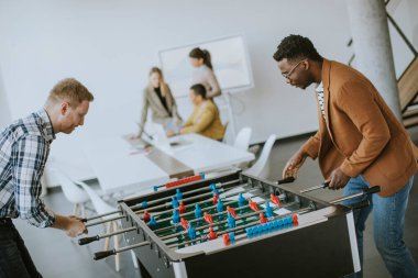 Genç, yakışıklı, çok ırklı iş adamları masa futbolu oynuyor ve ofiste dinleniyor.
