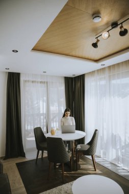 Genç bir kadın dizüstü bilgisayar kullanıyor ve odada sağlıklı portakal suyu içiyor.