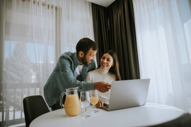 Genç çift oturma odasındaki masada dizüstü bilgisayar kullanıyor ve portakal suyu içiyor.
