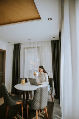 Genç bir kadın dizüstü bilgisayar kullanıyor ve odada sağlıklı portakal suyu içiyor.