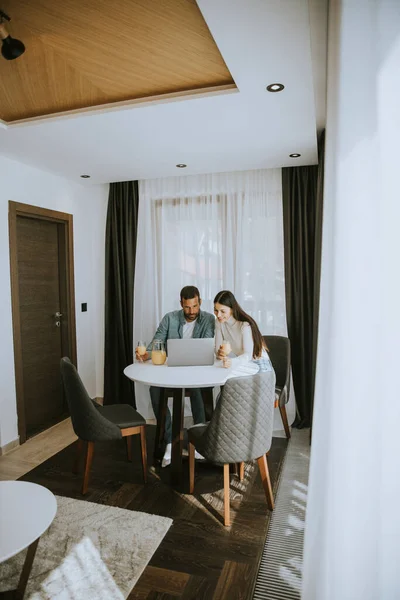 Genç çift oturma odasındaki masada dizüstü bilgisayar kullanıyor ve portakal suyu içiyor.