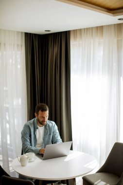 Yakışıklı genç adam dizüstü bilgisayar kullanıyor ve oturma odasında çay içiyor.