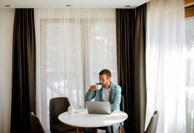 Yakışıklı genç adam dizüstü bilgisayar kullanıyor ve oturma odasında çay içiyor.