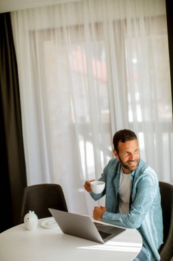 Yakışıklı genç adam dizüstü bilgisayar kullanıyor ve oturma odasında çay içiyor.