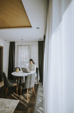 Genç bir kadın dizüstü bilgisayar kullanıyor ve odada sağlıklı portakal suyu içiyor.