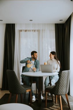 Genç çift oturma odasındaki masada dizüstü bilgisayar kullanıyor ve portakal suyu içiyor.
