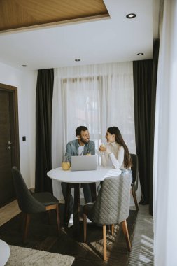 Genç çift oturma odasındaki masada dizüstü bilgisayar kullanıyor ve portakal suyu içiyor.