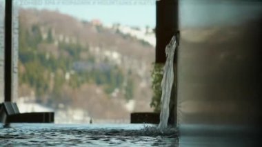 Su, arka planda kış dağları olan kapalı kaplıca havuzuna akar.