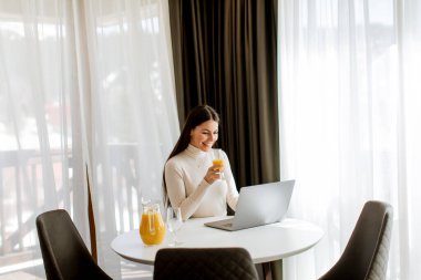 Genç bir kadın dizüstü bilgisayar kullanıyor ve odada sağlıklı portakal suyu içiyor.