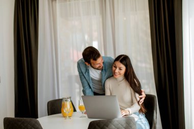Genç çift oturma odasındaki masada dizüstü bilgisayar kullanıyor ve portakal suyu içiyor.