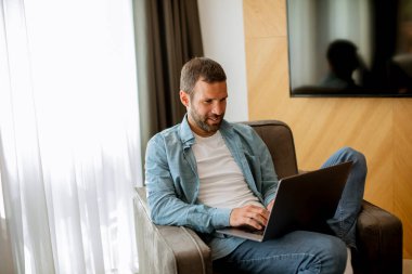 Yakışıklı genç adam oturma odasında dizüstü bilgisayar kullanıyor.