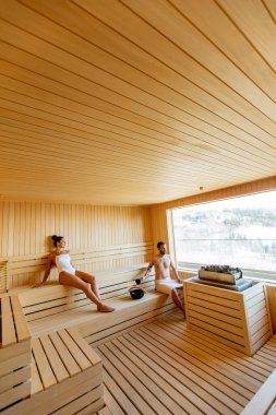 Yakışıklı genç çift saunada dinleniyor ve pencereden kış ormanlarını izliyorlar.