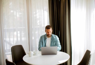 Yakışıklı genç adam dizüstü bilgisayar kullanıyor ve oturma odasında çay içiyor.