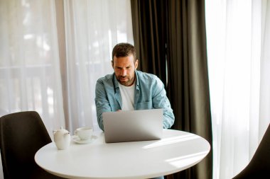 Yakışıklı genç adam dizüstü bilgisayar kullanıyor ve oturma odasında çay içiyor.