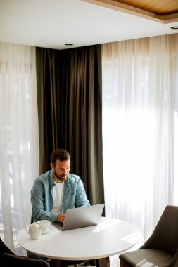Yakışıklı genç adam dizüstü bilgisayar kullanıyor ve oturma odasında çay içiyor.