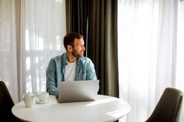 Yakışıklı genç adam dizüstü bilgisayar kullanıyor ve oturma odasında çay içiyor.