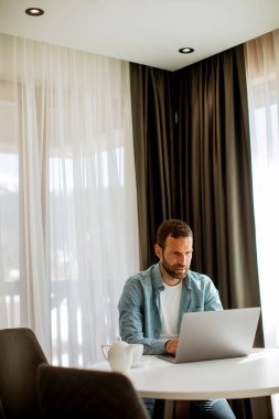 Yakışıklı genç adam dizüstü bilgisayar kullanıyor ve oturma odasında çay içiyor.