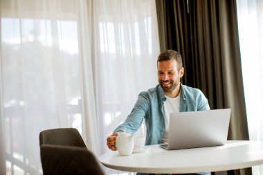 Yakışıklı genç adam dizüstü bilgisayar kullanıyor ve oturma odasında çay içiyor.